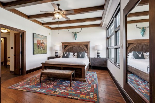 A rustic bedroom features a wood bed, colorful rug, animal art, and decorative antlers, with large windows and a ceiling fan.
