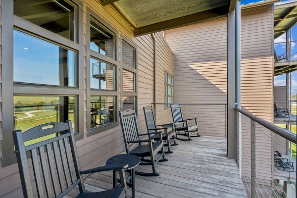 A wooden balcony features several black rocking chairs, offering a relaxing seating area. The setting overlooks a grassy landscape, ideal for leisure.