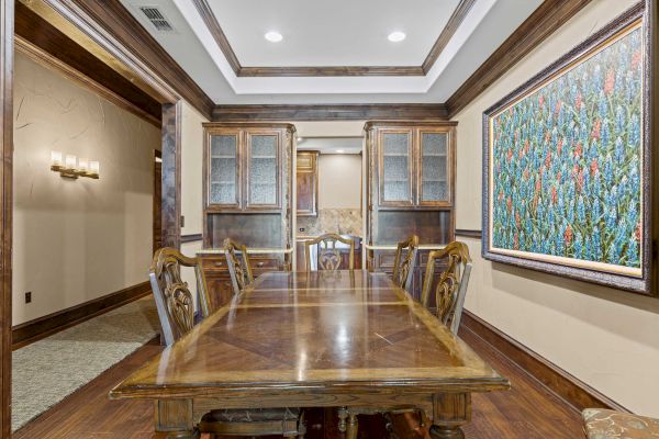 A dining room with a wooden table, chairs, cabinets, and a colorful painting on the wall. Recessed lighting above enhances the decor.