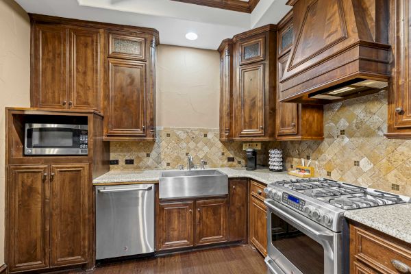 A warm-toned kitchen with wooden cabinets, a stainless steel stove, microwave, dishwasher, and a large sink, plus a tiled backsplash.