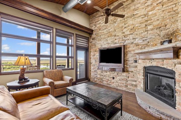 A cozy living room with a stone fireplace, leather furniture, large windows, and a flat-screen TV mounted on the wall.