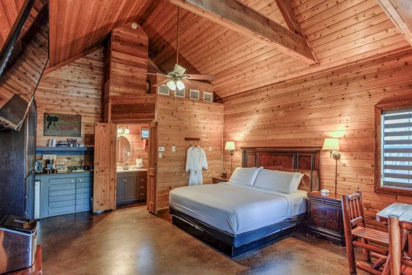 A cozy wooden cabin interior with a bed, wooden furniture, a kitchenette, and a bathroom, featuring warm lighting throughout.