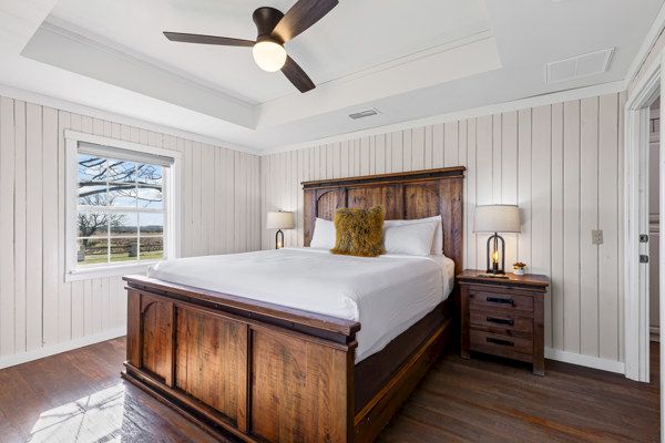 A cozy bedroom with a wooden bed, nightstands, lamps, and a ceiling fan, featuring white paneled walls and a window view.
