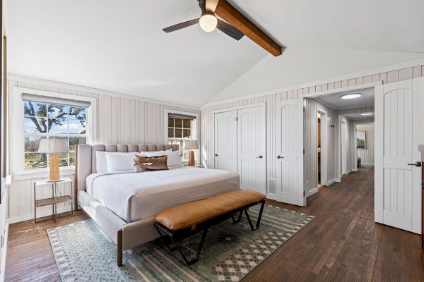 A cozy bedroom features a bed with a tufted headboard, a bench, wooden flooring, and a vaulted ceiling with a fan.