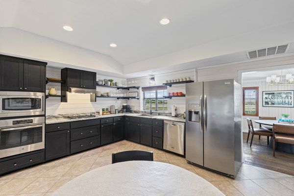 A modern kitchen with dark cabinets, stainless steel appliances, marble countertops, and a dining area visible in the background.