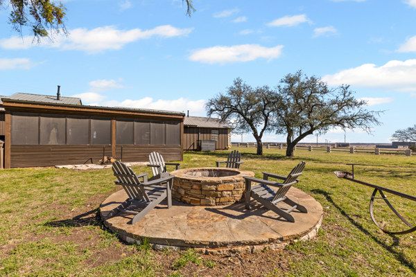 A stone fire pit with chairs on a patio, near a wooden house and trees under a blue sky with clouds.