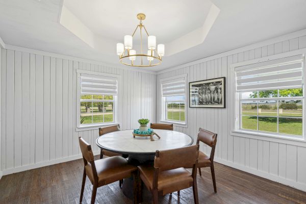 A dining room with white shiplap walls, a round table with four chairs, two windows, and a chandelier above.