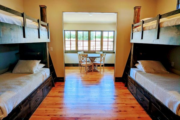 The image shows a room with two sets of bunk beds, hardwood floors, and a table with chairs by a window in the background.