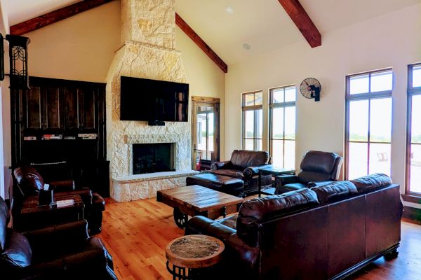 A cozy living room with wooden floors, leather sofas, a stone fireplace, large windows, and a unique ceiling fan with beveled beams.