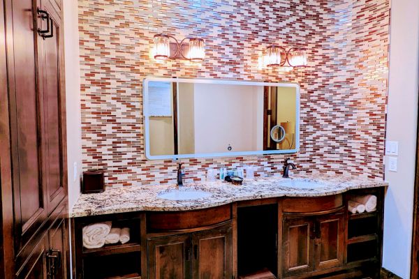 A bathroom with a patterned tile wall, double sink vanity, large mirror, and wooden cabinetry, creating a warm and elegant atmosphere.
