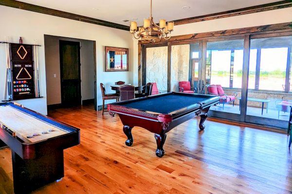 The image shows a game room with a billiards table, shuffleboard, chairs, a chandelier, and large windows.