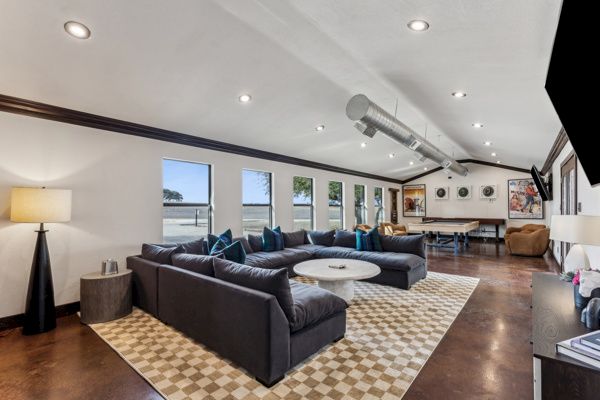 A modern living room with a large sectional sofa, round table, pool table, and wall-mounted TV. Neutral tones and decorative elements.