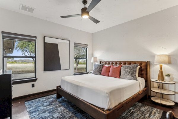 A cozy bedroom with a large bed, decorative pillows, a rug, two windows, abstract wall art, a ceiling fan, and a lamp on a side table.