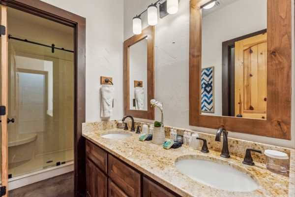 A bathroom with a double sink vanity, granite countertop, and mirror. There's a glass shower door and wooden accents throughout the space.
