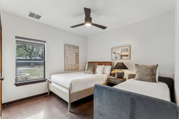 A bedroom with two beds, a ceiling fan, a window, framed art, and a nightstand with a lamp and flower vase.