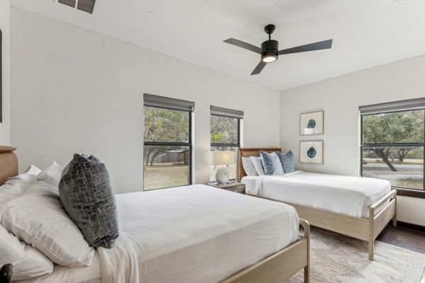 The image shows a bedroom with two beds, decorative pillows, a ceiling fan, artwork, and windows overlooking trees.