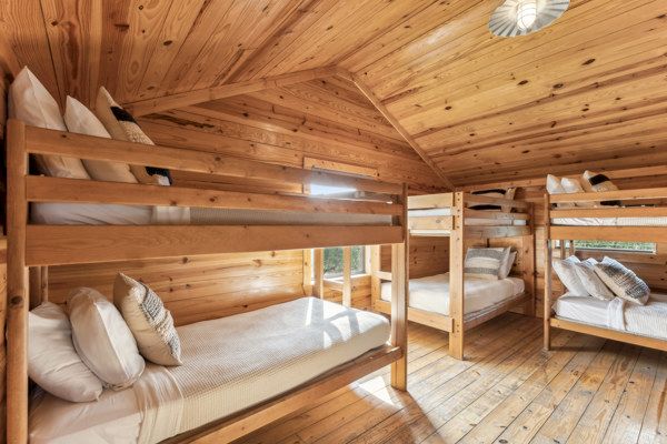 The image shows a cozy wooden room with two sets of bunk beds, each neatly made with pillows and blankets.