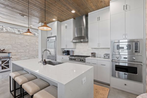 Modern kitchen with white cabinets, island with stools, stainless steel appliances, pendant lights, and wooden ceiling.