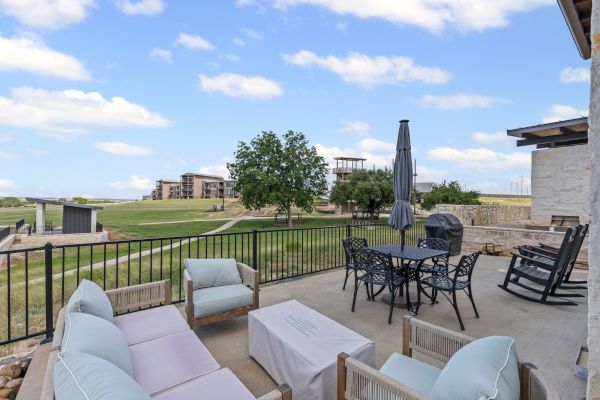 A patio with seating, a table, grill, and umbrella overlooks a scenic grassy area with buildings in the distance under a blue sky.