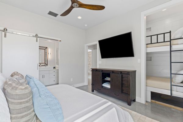 A modern bedroom with a bed, wall-mounted TV, ceiling fan, bunk beds, and a bathroom visible through a sliding door.