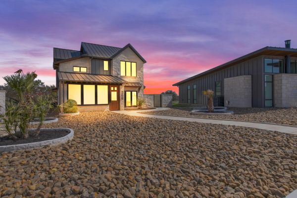 A modern house with illuminated windows sits under a vibrant sunset sky, surrounded by a stone-filled landscape.