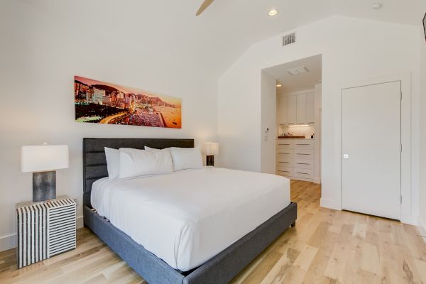 A modern bedroom features a large bed, two lamps, a vibrant cityscape artwork above the bed, and light wood flooring.