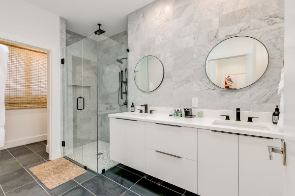 Modern bathroom with a glass shower, dual sinks, and circular mirrors. The design features white cabinetry and marble accents.