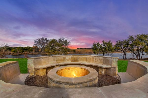 An outdoor fire pit with stone seating overlooks a serene lake at sunset, surrounded by trees under a colorful sky.