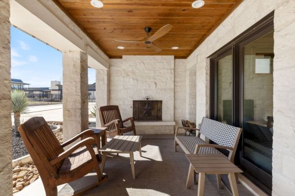 Outdoor patio with wooden chairs, a bench, small tables, a ceiling fan, and a fireplace against a stone wall, offering a cozy setting.