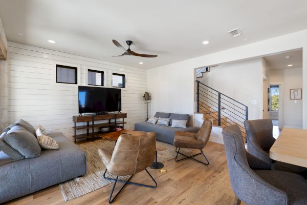 A modern living room with sofas, armchairs, a TV, ceiling fan, and staircase, featuring wooden flooring and a minimalist design aesthetic.