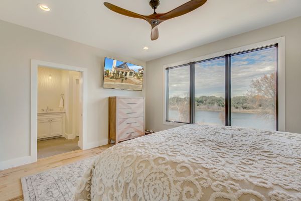 A bedroom with a large bed, ceiling fan, wall-mounted TV, dresser, and windows with a scenic view; an open door leads to a bathroom.