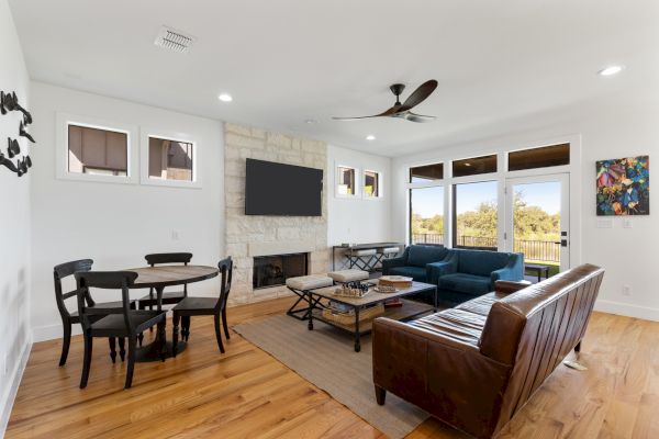 A cozy living room features a blue couch, brown leather sofa, small dining table, wall-mounted TV, and large windows with a scenic view.