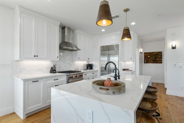 A modern kitchen with white cabinets, marble island, bar stools, pendant lights, and stainless steel appliances. Warm wooden floor enhances it.