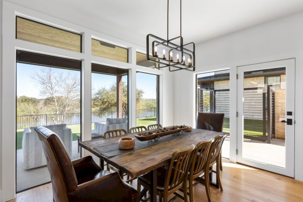 A dining room with a wooden table, multiple chairs, large windows, and a pendant light fixture overlooking a patio.