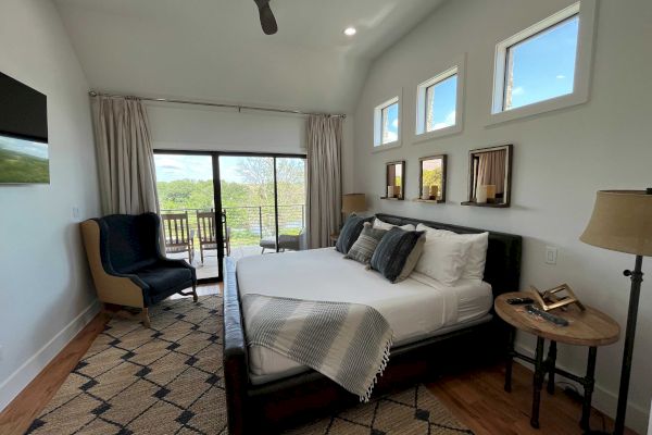A modern bedroom with a bed, armchair, lamps, and decor. There’s a large window with curtains and an outside view.