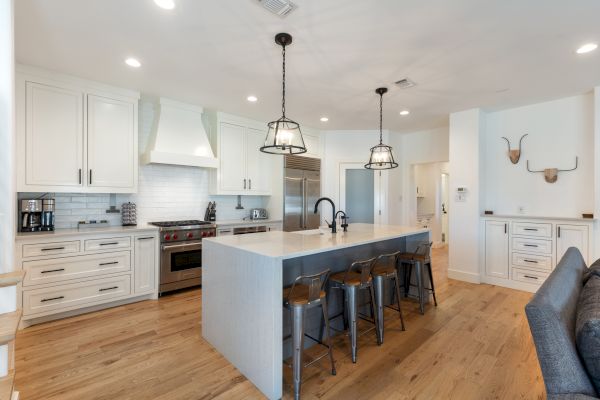A modern kitchen with an island, bar stools, pendant lights, and stainless steel appliances on wooden flooring and white cabinets.