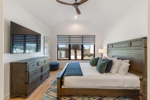A modern bedroom with a wooden bed, dresser, TV, teal accents, a window, ceiling fan, and a patterned rug, in a clean design style.