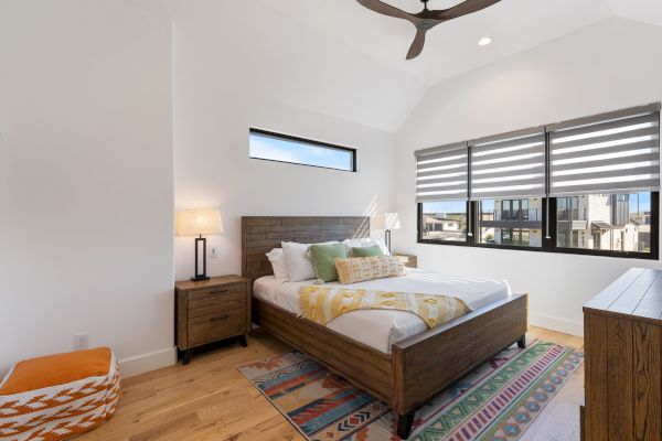 A modern bedroom with a wooden bed, nightstand, and dresser, featuring patterned rugs and large windows with blinds.