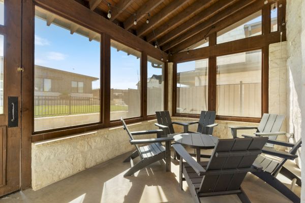 The image shows a sunlit patio with wooden chairs and a table, enclosed by large glass windows, and a view of a fenced yard outside.