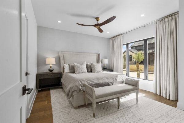 A modern bedroom with a bed, bench, nightstand, and lamp, featuring a ceiling fan and large window with curtains, in a bright setting.