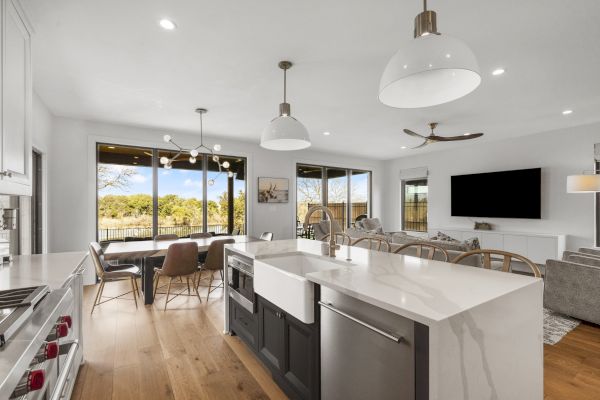 A modern kitchen and living area with an island, light wood floors, and a view outside through large windows, featuring a TV and ceiling fans.