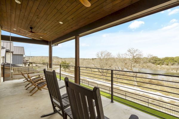 A covered patio features wooden rocking chairs overlooking a scenic view with trees, grass, and a river under a blue sky with clouds.
