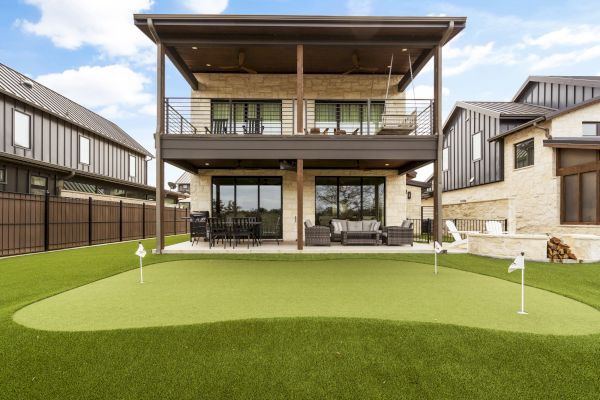 A modern house with a balcony overlooks a backyard putting green, surrounded by a neatly manicured lawn, perfect for golf practice.