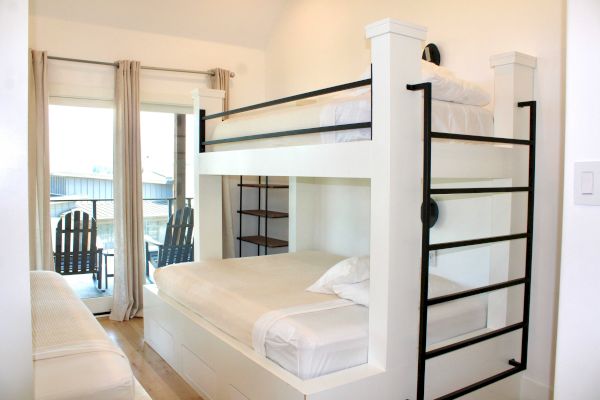 The image shows a cozy bedroom with a bunk bed, white bedding, and a small balcony featuring two chairs and an outdoor view.