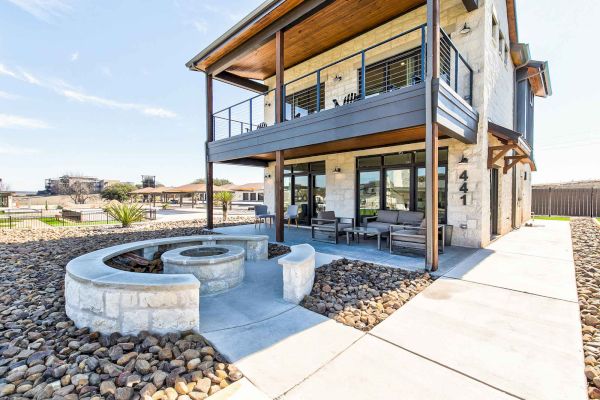 The image shows a modern two-story house with a balcony, stone façade, outdoor seating, and a round stone planter or fire pit area.