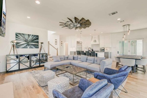 The image shows a modern living room with a large sectional sofa, a kitchen in the background, stylish decor, and wooden flooring.