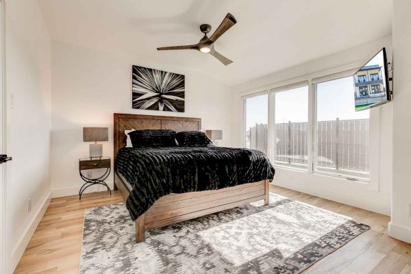 A modern bedroom with a bed, black bedding, two side tables, a ceiling fan, large windows, and a wall-mounted TV.