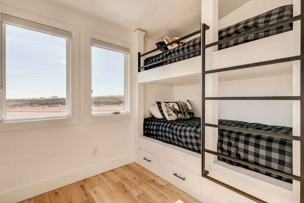 A room features two bunk beds with plaid bedding, a ladder, two windows, and wooden flooring.
