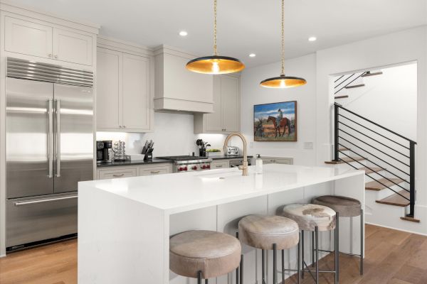 A modern kitchen features a large island with stools, stainless steel appliances, and vibrant pendant lights, next to a staircase.