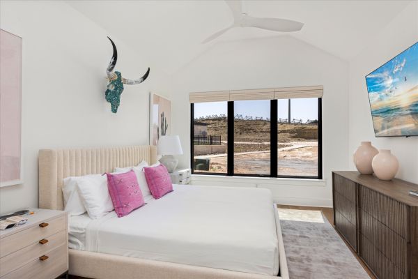 A stylish bedroom with a large bed, pink pillows, artwork, a window view, and a wall-mounted TV for modern comfort and design appeal.